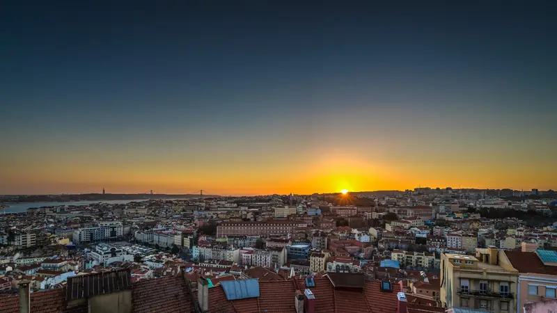 Sunset over the red rooftops of Lisbon seen from Miradouro da Senhora do Monte, with the Tagus river and the 25 de Abril bridge on the horizon