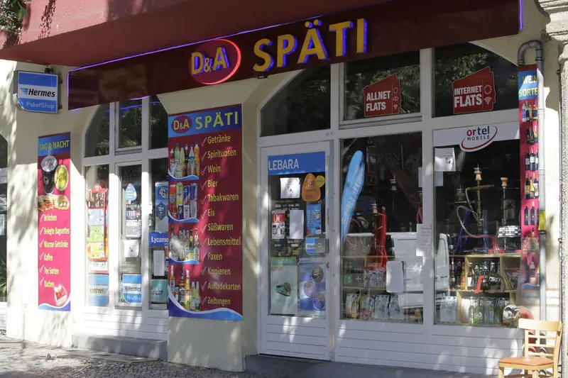 Storefront of a Späti late-night shop in Berlin with a red and yellow sign and window posters listing drinks, snacks and coffee