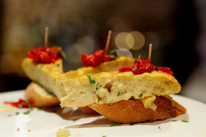Slices of Spanish potato omelette on bread, topped with roasted red pepper and held with toothpicks, on a white plate in a Barcelona bar