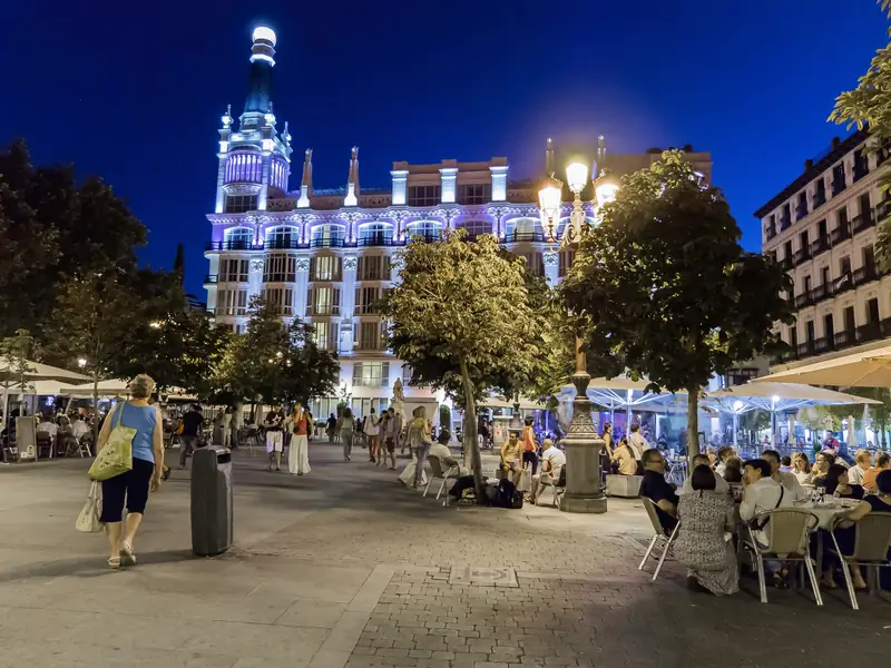 Plaza de Santa Ana in Madrid at night, with people eating at terrace tables under umbrellas in front of a lit white hotel building with a tower