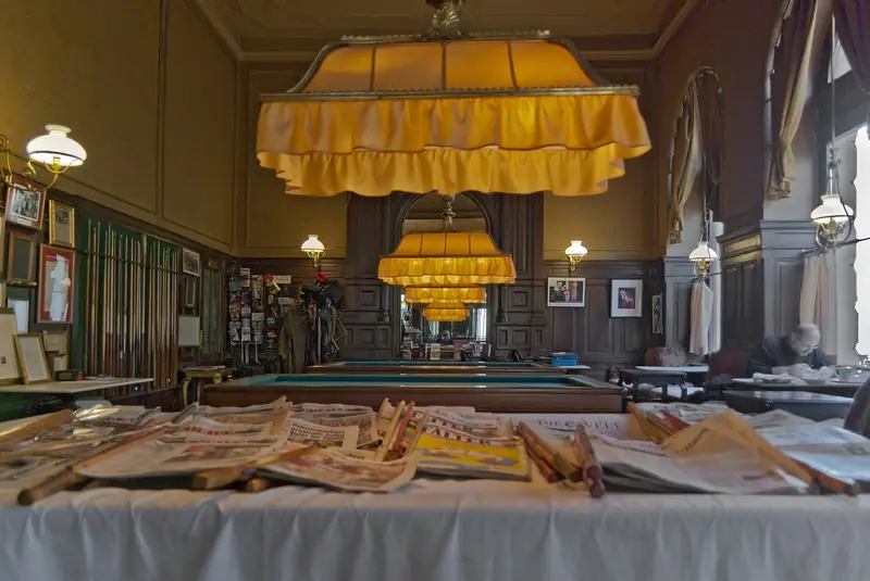 Wooden newspaper holders and daily papers on a table in Café Sperl, with billiard tables and fringed yellow lamps behind